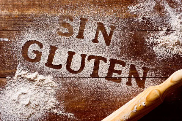 high-angle shot of a wooden table sprinkled with a gluten free flour where you can read the text sin gluten, gluten free written in spanish, next to a rolling pin