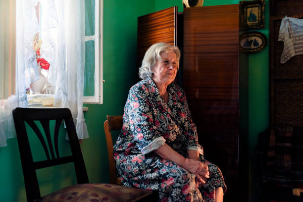 Old lonely woman sitting near the window in her house. Photo of One elderly woman looks sadly while sitting next to window and looking away. Elderly woman with memory problem spending time in her rustic home  during the day. Old woman sitting on chair with a worried expression on her face.