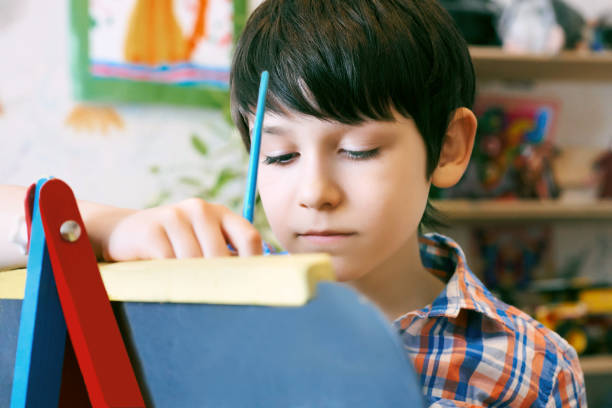 istock Child standingnext to the easel. Kid boy learn paint by brush in class school. Kindergarten interior on background. Boy is getting ready to become an artist 961345018