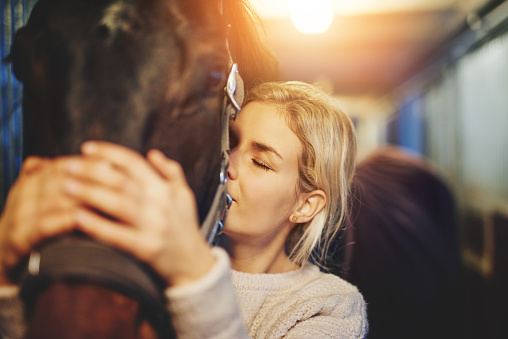 Smiling young woman standing in a stable affectionately hugging her chestnut horse before going for a ride