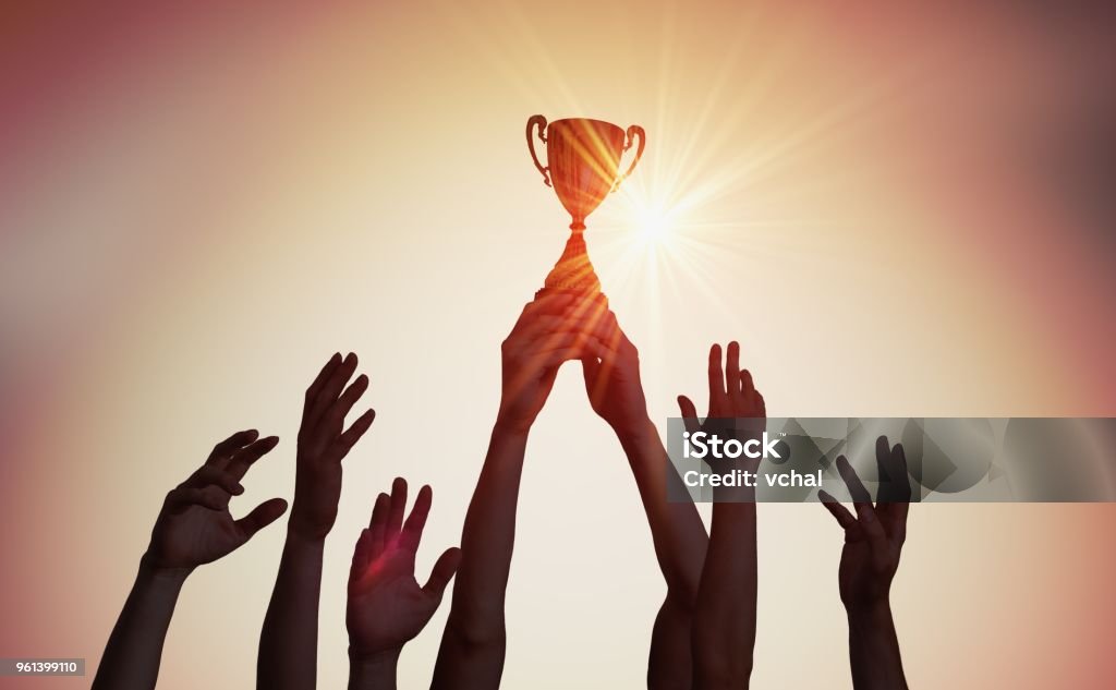 Equipa vencedora está segurando o troféu nas mãos. Silhuetas de muitas mãos no pôr do sol. - Foto de stock de Vencer royalty-free