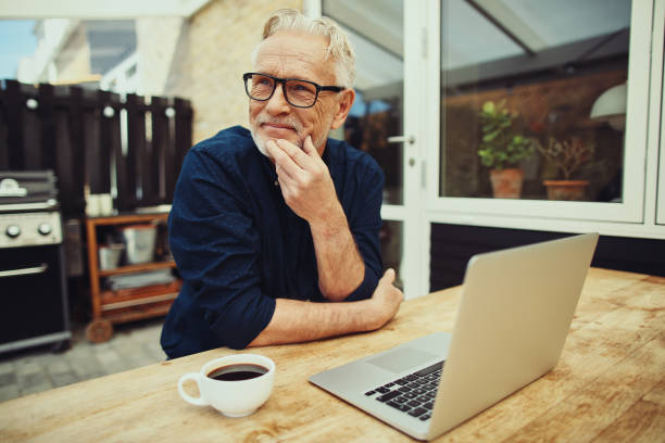 senior man sitting outside drinking coffee and using a laptop - computer thinking men people imagens e fotografias de stock