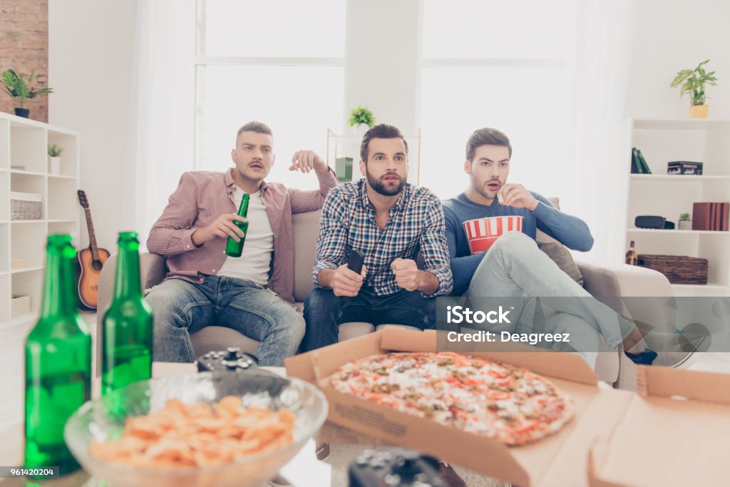 Retrato de chicos atractivos, con estilo con peinado moderno viendo futbol de palabra con excitada expresión beber bebidas de alcohol, comer pizza, patatas fritas, snack, sentado en la sala de estar - Foto de stock de Fútbol libre de derechos