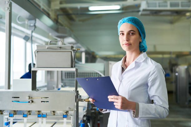 istock Serious attractive female quality engineer in sterile clothes holding clipboard with record and looking at camera while standing at food manufacturing machine in factory 961452590
