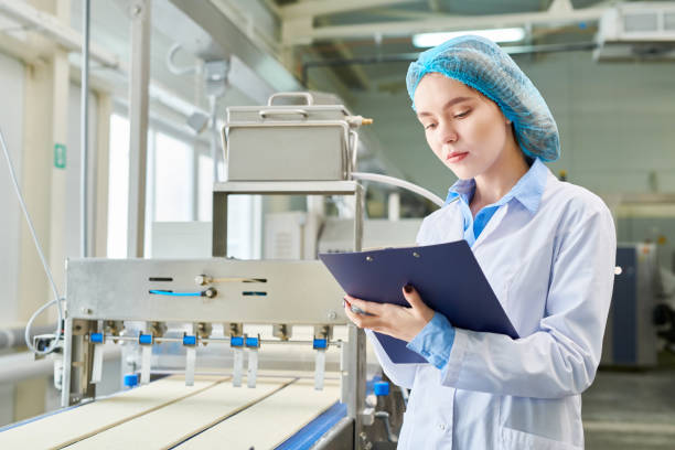 istock Serious concentrated female food engineer in sterile cap and coat keeping record while estimating conveyor line producing pastry dough in factory 961453242