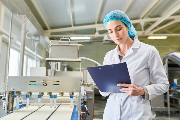 istock Serious concentrated attractive female quality control specialist in protective cap and lab coat writing information about dough rolling machine in clipboard at plan 961453362