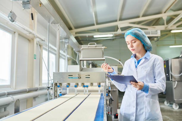 istock Concentrated beautiful young female food manufacturing engineer in sterile clothes analyzing data while examining industrial machine at factory 961453376