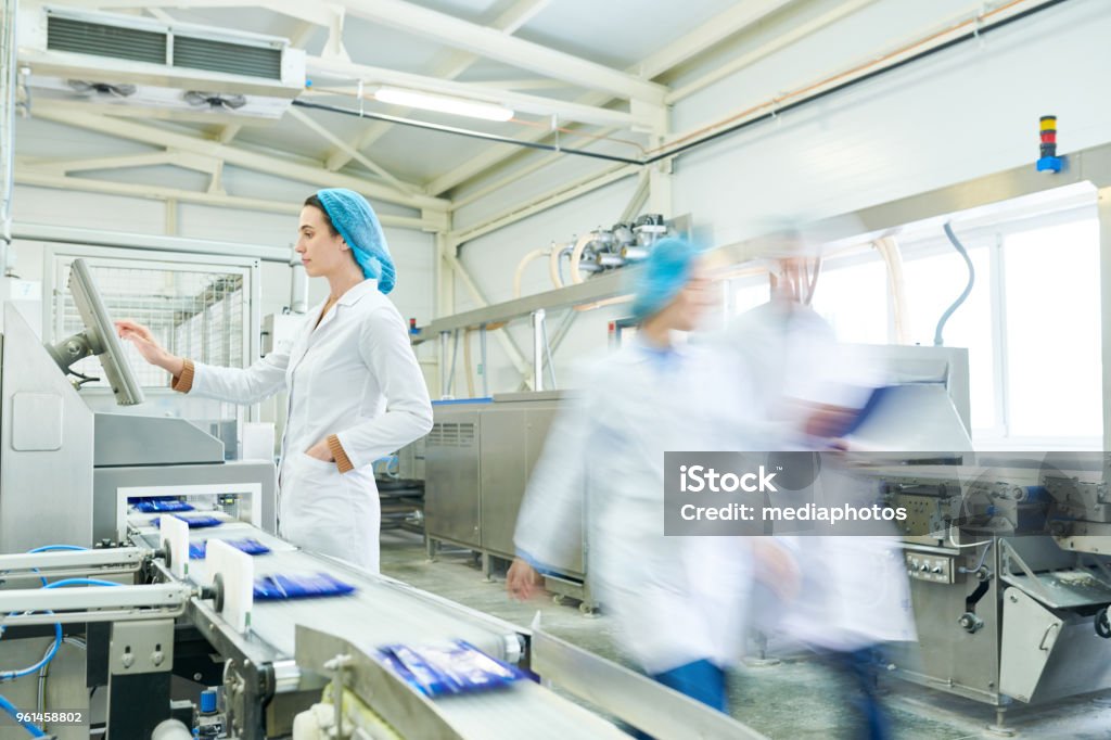 Busy female worker in sterile clothes choosing program on touch screen while operating manufacturing machine producing packaged food, blurred motion of technologists Food production line staff Food Processing Plant Stock Photo