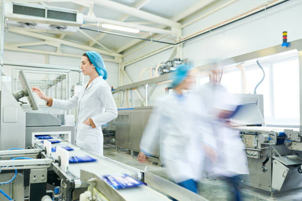 Busy female worker in sterile clothes choosing program on touch screen while operating manufacturing machine producing packaged food, blurred motion of technologists Food production line staff food processing plant stock pictures, royalty-free photos & images