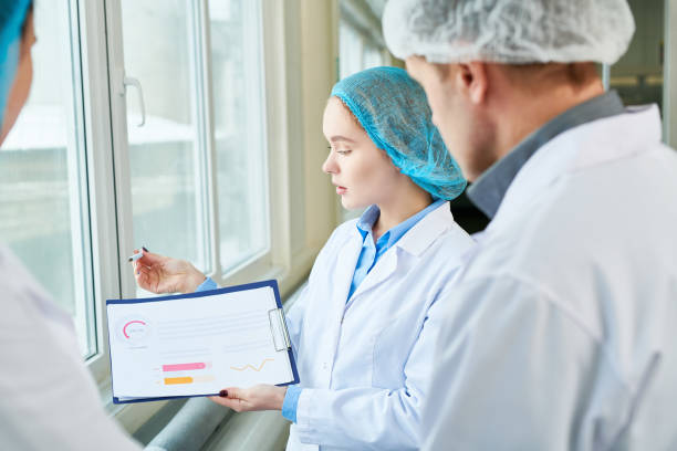 Serious confident female food technologist holding chart and presenting statistic to colleagues while they discussing consumption issues in factory