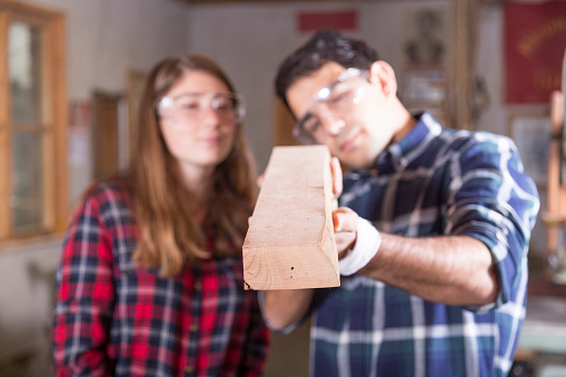 Young male carpentry manual worker manufacturing at workshop. Wears shirt and safety goggles on head. Tools on shelves and wooden material on background. Focus on foreground.