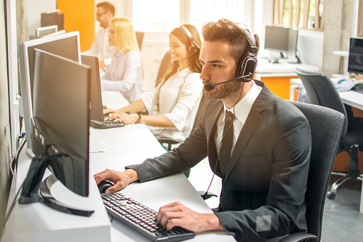 Friendly handsome man in suit with headset working with colleagues in call center