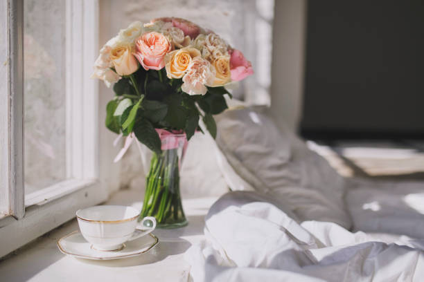 A charming large bouquet of roses and a white tea cup is on the windowsill next to the bed. stock photo