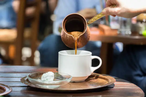 Woman hand pouring greek coffee from a copper pot on a white cup placed on a tray with loukumi