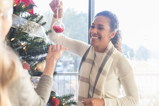 Two mid adult women enjoy sharing memories with each ornament they hang together on a Christmas tree.