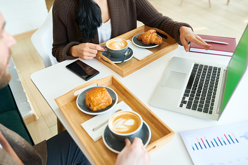 High angle crop shot of two business people, man and woman, enjoying coffee break while working in cafe, focus on coffee cups and croissants on wooden trays by laptop with green screen