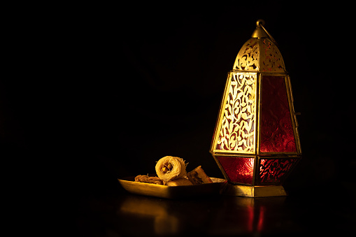 Traditional ornamental arabic lantern with a burning candle in dark studio setting with traditional arabic sweets. Festivel greeting card for Ramadan Kareem and Ramadan Mubarak. Dubai, UAE.
