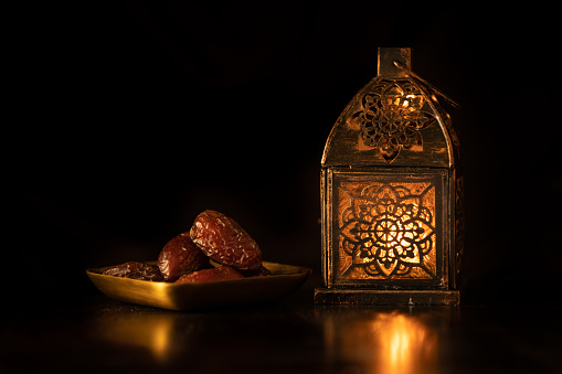 Traditional ornamental arabic lantern with a burning candle in dark studio setting with majool dates. Festivel greeting card for Ramadan Kareem and Ramadan Mubarak. Dubai, UAE.