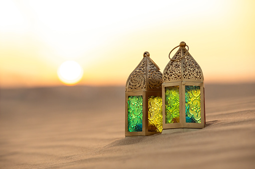 Traditional ornamental arabic lantern with a burning candle in desert during sunset. Festivel greeting card for Ramadan Kareem and Ramadan Mubarak. Dubai, UAE.