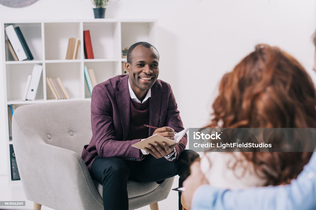 Sorridente psichiatra afroamericano che parla con giovane coppia - Foto stock royalty-free di Psicoterapeuta