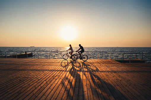 Couple of young hipsters cycling together at the beach at sunrise sky at wooden deck summer time.