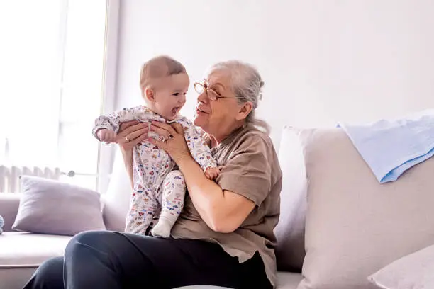 Grandmother holding little grandson in the room at home. Senior woman hold little baby cute smiling. Happy grandmother with her grandchild in home.