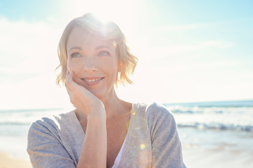 Beautiful mature woman portrait on the beach. She is happy and smiling with the sun and sea behind her. She looks relaxed and could be on vacation.