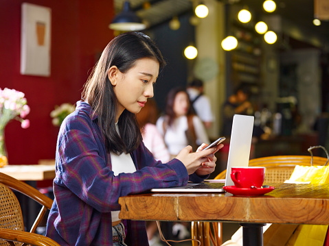 young asian woman sitting working in coffee shop using mobile phone and laptop computer.
