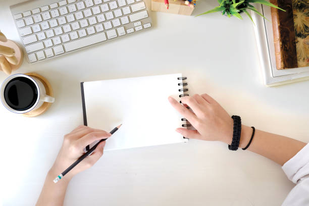 istock Top view female hand writing blank notebook on white office desk and computer with office supplies. work space concept 961648564