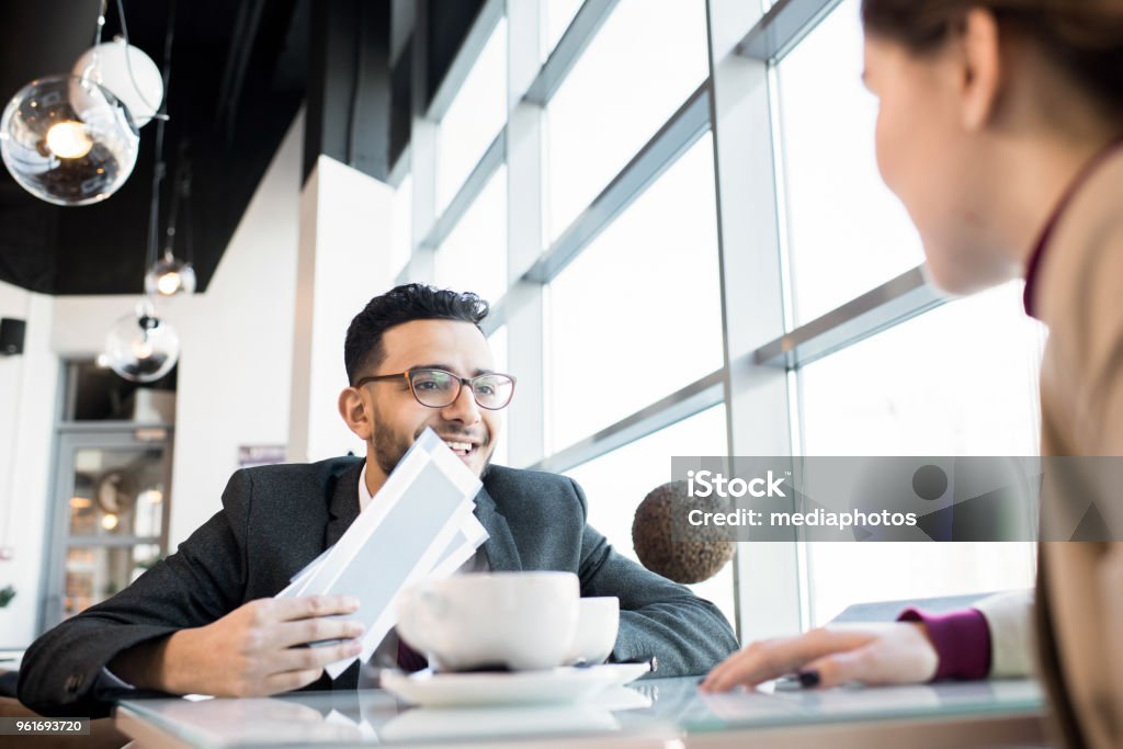 Positive successful young Arabian businessman excited about business trip and showing airline tickets to colleague in cafe Boss sending best manager to travel for work Business Stock Photo