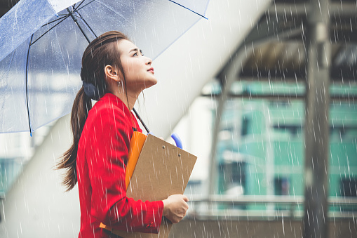 Young asian businesswoman holding business documents and umbrella walking under raining in the city