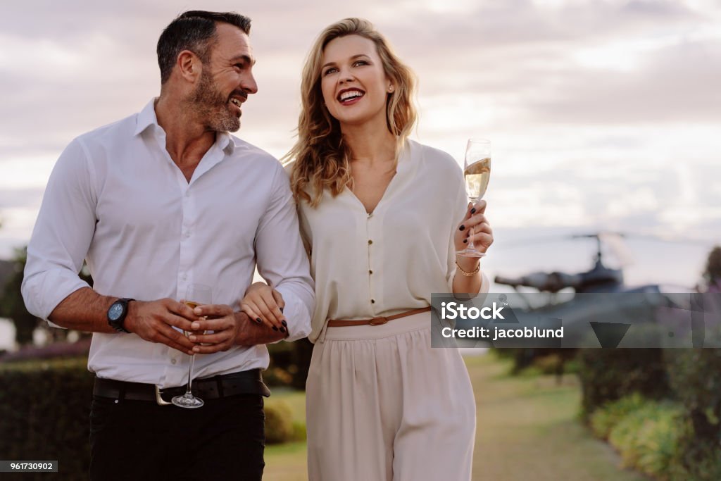 Smiling couple walking outdoors Smiling couple walking outdoors holding a glass of wine. Caucasian man and woman with a drinks walking together with a helicopter in background. Couple - Relationship Stock Photo