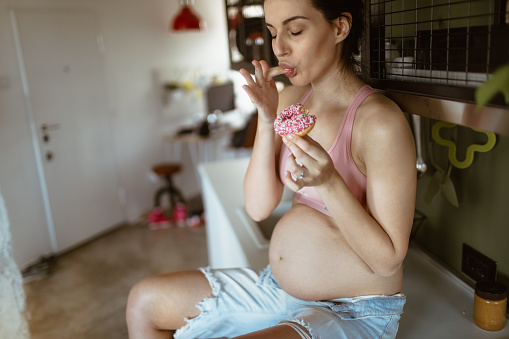 Portrait of a future mother having hard times to maintain healthy lifestyle while pregnant - and eating donuts
