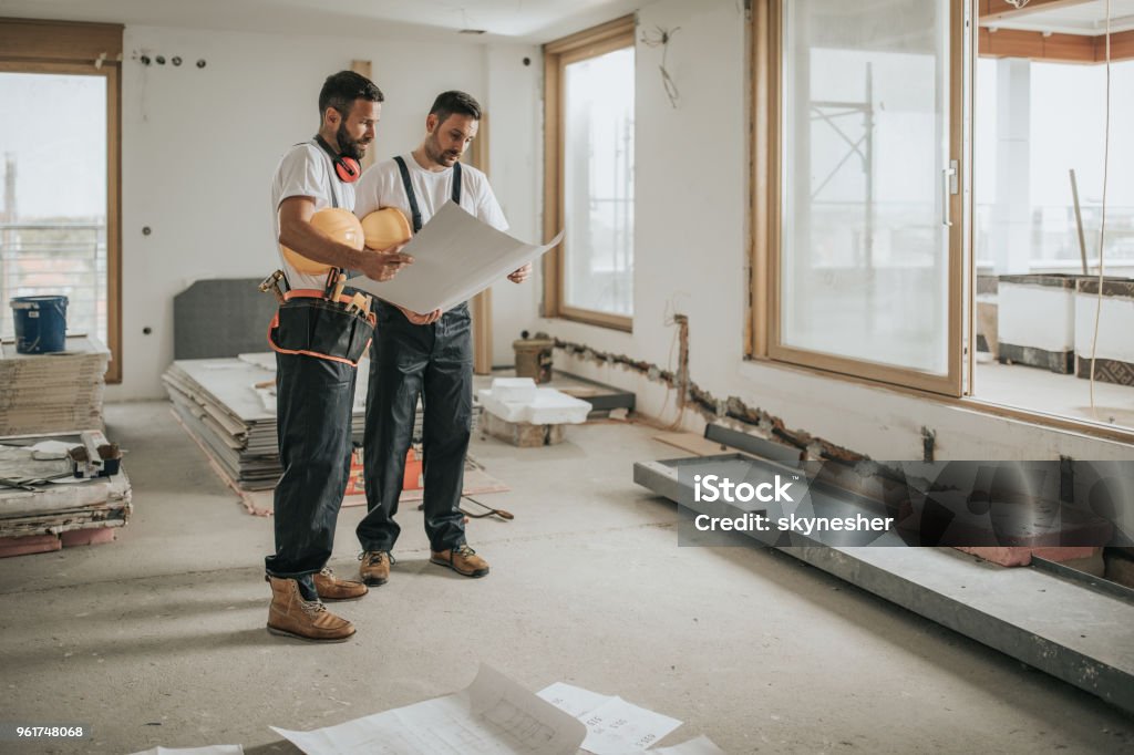 Full length of construction workers analyzing blueprints in the apartment. Young manual workers standing at construction site and examining blueprints. Construction Industry Stock Photo