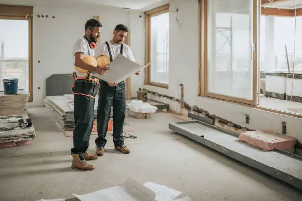 Young manual workers standing at construction site and examining blueprints.