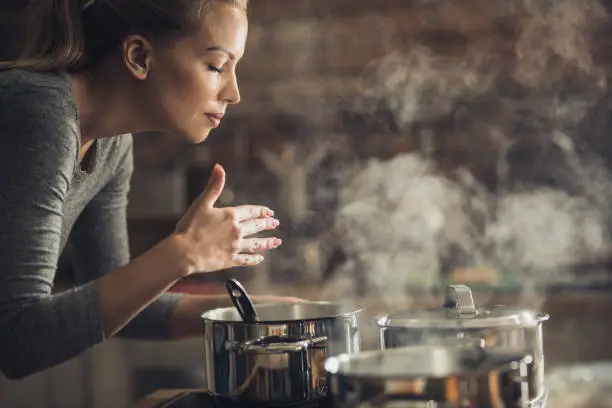 Young woman smelling soup that she is preparing for lunch.