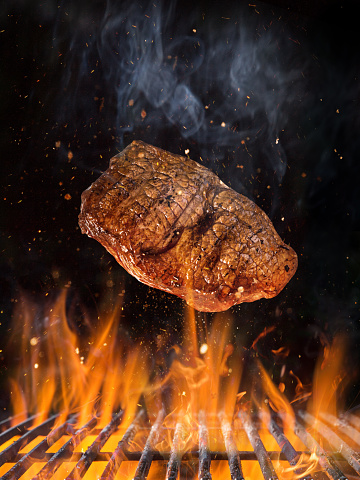 Tasty beef steak flying above cast iron grate with fire flames. Freeze motion barbecue concept.