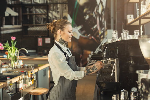 Young tattooed barista making coffee in professional coffee machine. Woman preparing beverage. Small business and professional coffee brewing concept