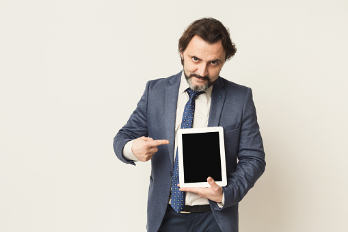 Bearded man holding digital tablet with blank screen, showing it, white studio background, copy space