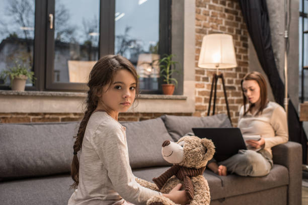 istock selective focus of upset kid with teddy bear looking at camera while mother working on laptop on sofa at home 961845156