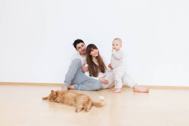 Happy weekend on young family loving parents looking at their adorable baby next to red cat sitting on the floor in white pajamas