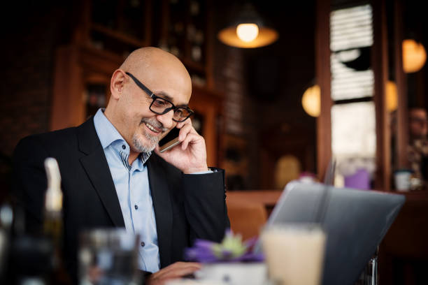 istock Mature businessman is smiling during his phone call while using laptop and sitting at the table of a coffee shop. 961853710