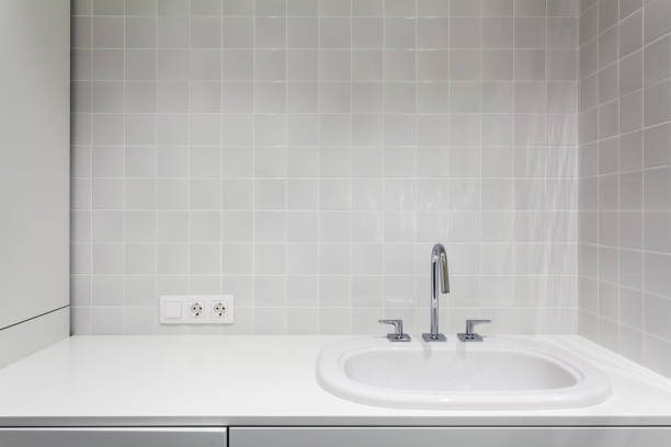 A modern bathroom design featuring a simple white ceramic sink with a chrome faucet, mounted beneath a mirror and glass shelf. The tiled wall includes a power outlet, towel ring, and minimal accessories, highlighting a clean and functional layout.