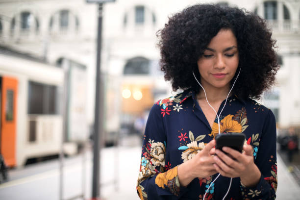 Woman at the train station using mobile phone. Smiling African American woman with curly hair using mobile phone at the train station. podcasting stock pictures, royalty-free photos & images