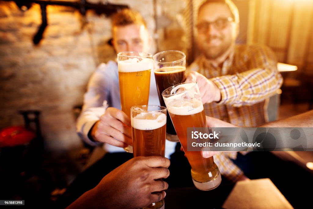 A group of young men clinking glasses with a beer in the sunny pub. Beer - Alcohol Stock Photo