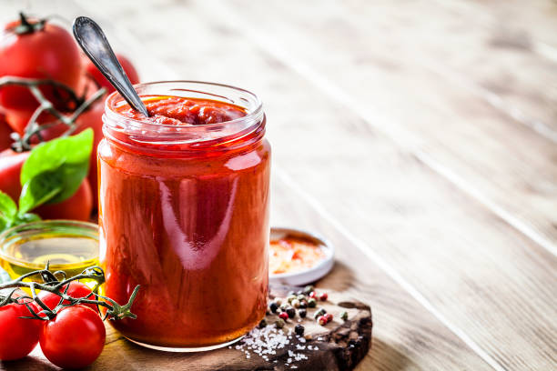 Tomato sauce jar Preparing tomato sauce at home. Front view of an open glass jar filled with prepared tomato sauce shot on wooden kitchen table with ingredients. The composition is at the left of an horizontal frame leaving useful copy space for text and/or logo at the right. Ingredients for preparing tomato sauce are fresh ripe tomatoes, basil, olive oil, garlic, salt and pepper. Predominant color is red. Low key DSRL studio photo taken with Canon EOS 5D Mk II and Canon EF 100mm f/2.8L Macro IS USM preserved food stock pictures, royalty-free photos & images