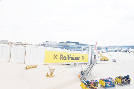 Several ground support vehicles on a tarmac servicing a number of airplanes at Vienna, Austria. Dollies transporting baggage and ad board (Raiffeisen Bank) on the elevated walkway