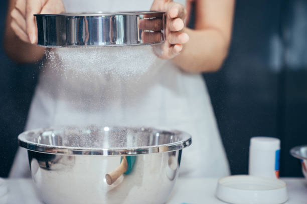 uma mulher de peneirar a farinha em uma tigela de aço inoxidável. menina é peneirar a farinha para fazer a padaria na cozinha no fim de semana. - sifting - fotografias e filmes do acervo