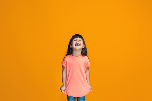 Happy teen girl standing, smiling isolated on trendy orange studio background. Beautiful female half-length portrait. Human emotions, facial expression concept.