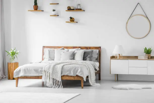 Photo of Comfortable big wooden framed bed with linen, pillows and blanket, nightstand beside and round mirror hanging on a white wall in a bright bedroom interior. Real photo.